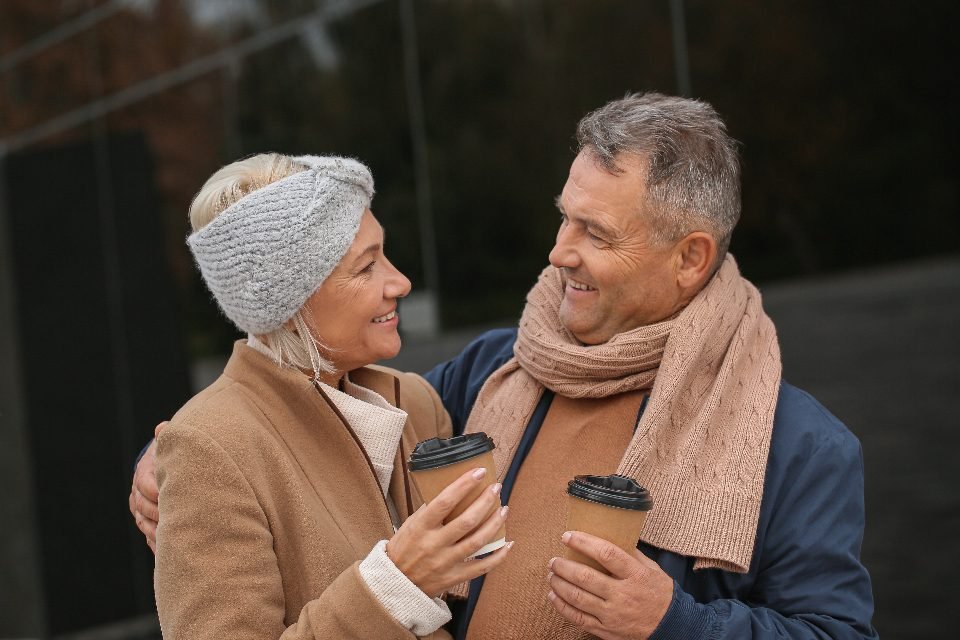 Smiling mature couple in winter scarf and headband share coffee outdoors, illustrating 45 plus dating sites