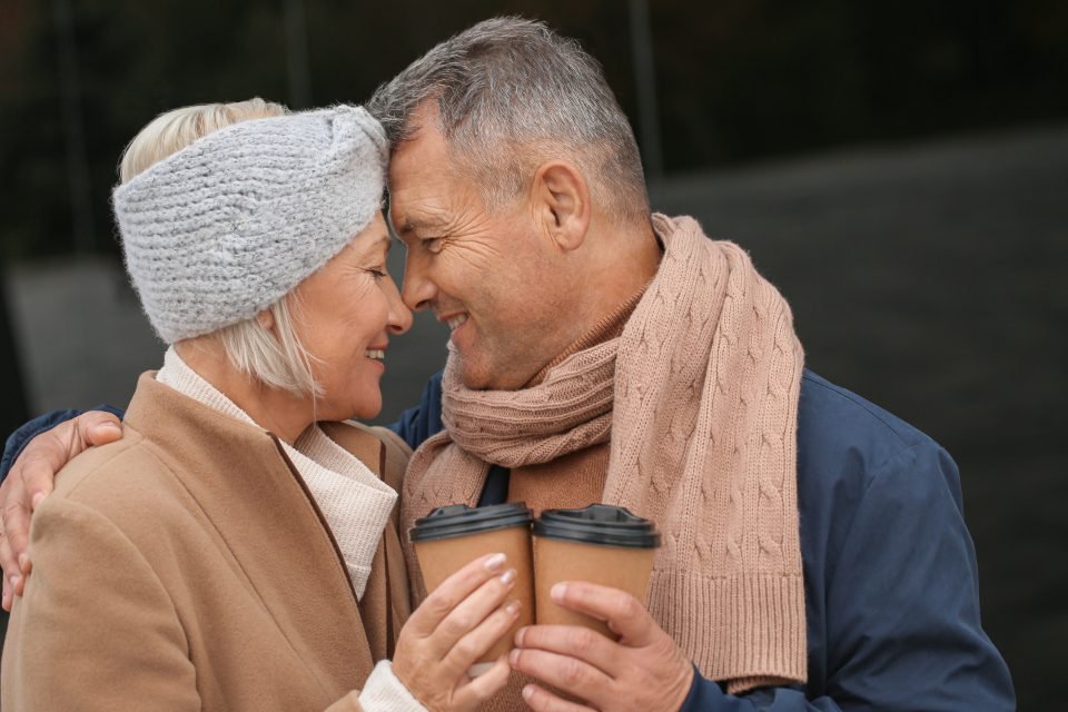 Smiling middle-aged couple touching foreheads outdoors, each holding a coffee cup, for 50 plus singles dating