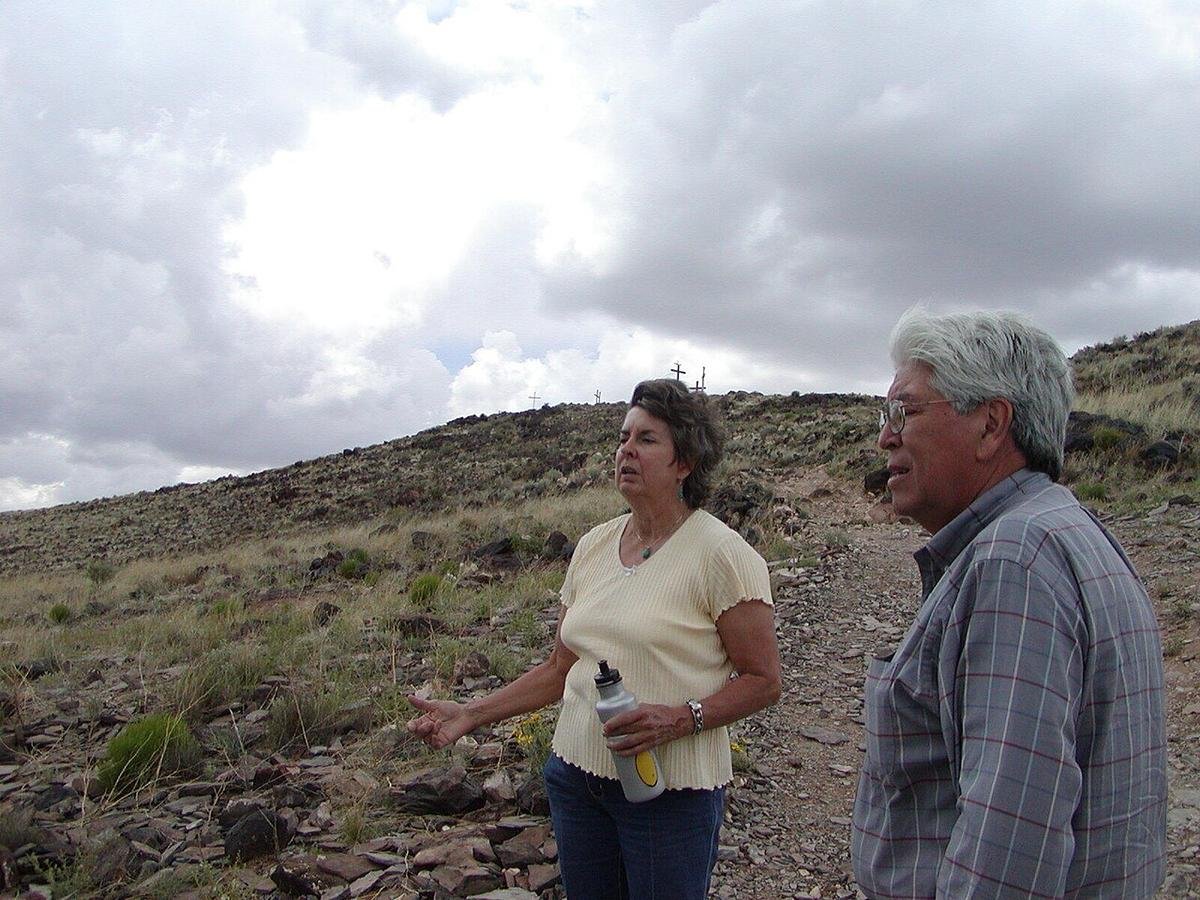 Older woman in cream sweater with water bottle gestures beside gray-haired man in plaid shirt on a dry rocky hillside.