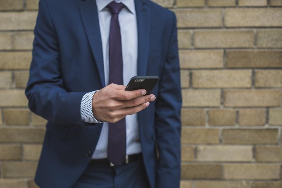 Man in a navy suit and purple tie typing a message on a smartphone against a brick wall