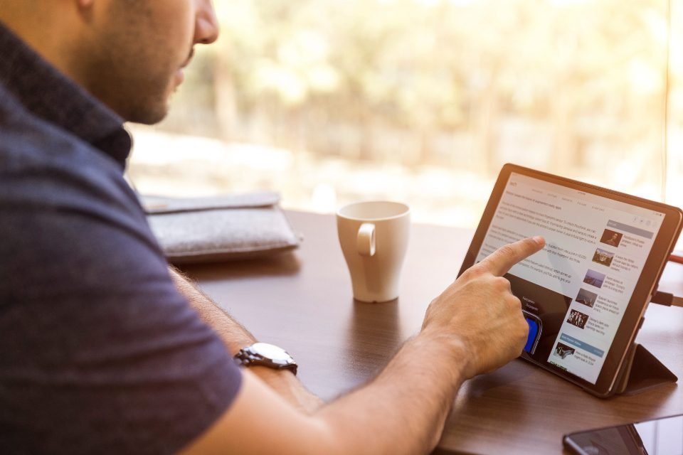 Man in a dark shirt pointing at a tablet showing an article, with a coffee mug and wristwatch on a wooden table by a window.
