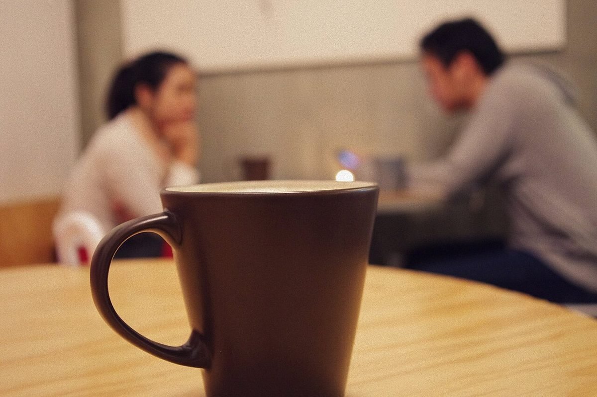 Blurred woman and man talking in a café; a brown coffee mug sits in sharp focus on a wooden table in front.