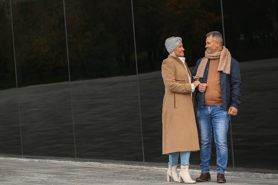 Smiling couple in winter coats and scarves standing face to face beside a dark glass wall outdoors.