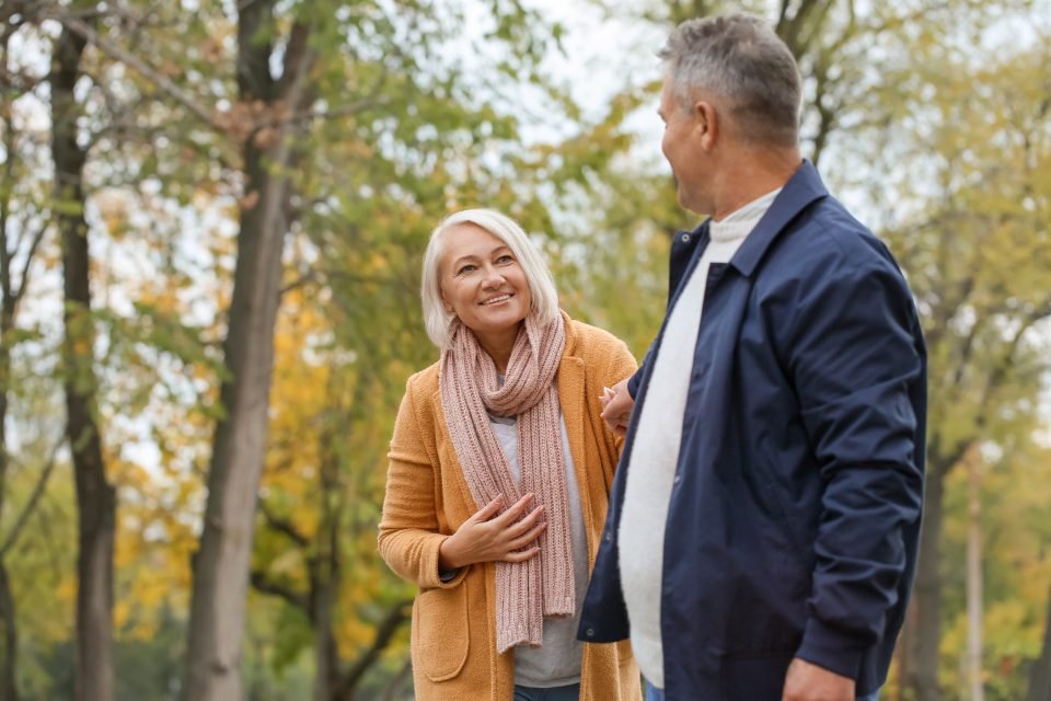 Smiling older woman in mustard cardigan and pink scarf chats with a gray-haired man in navy jacket in an autumn park.